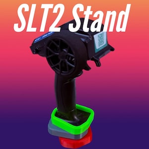 Op de afbeelding: Een zwarte afstandsbediening met een groene, grijze en rode standaard. De tekst "SLT2 Stand" staat in het wit bovenaan de afbeelding. De afstandsbediening heeft een zwart wiel en een klein scherm.
