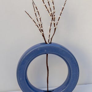 May include: A blue ceramic vase with a circular design, holding several brown stems with small white buds. The vase has a smooth, matte finish and sits on a white surface. The stems are arranged to create a simple, elegant floral display.