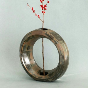 May include: A unique, circular ceramic vase with a brown and bronze glaze. A slender branch with red berries is placed in the vase. The vase has a modern, minimalist design, suitable for displaying flowers or decorative branches.
