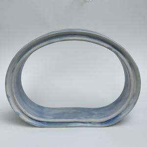 May include: A ceramic sculpture with a rounded, oval shape. The piece is primarily light blue with subtle gray accents, creating a soft, textured appearance. The design is minimalist and modern, suitable for home decor.