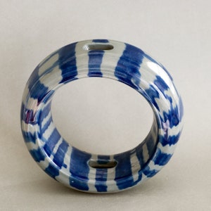 May include: A ceramic ring-shaped vase with a blue and white striped pattern. The vase has two rectangular openings on opposite sides. The design features alternating bands of blue and white, creating a visually appealing aesthetic. The vase is a decorative item.