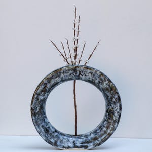May include: A ceramic ring vase with a speckled, textured surface in shades of gray, brown, and white. The vase holds a single, slender brown stem with delicate white buds, creating a minimalist floral arrangement. The vase is a unique decorative piece.