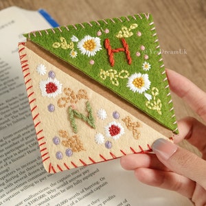 Puede incluir: Marcapáginas de fieltro bordado con diseños florales e iniciales. El marcapáginas presenta una combinación de colores verde y beige, con costuras rojas y flores bordadas. La inicial "H" es roja y "N" es verde.