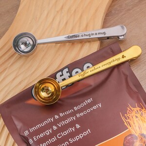 May include: Two gold-tone coffee measuring spoons with engraved sayings. The first spoon says "Coffee: a hug in a mug." The second spoon says "Coffee solves everything."