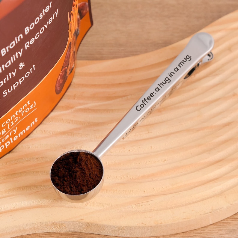 May include: A silver measuring spoon with a clip on the handle. The spoon is filled with dark brown coffee grounds. The spoon has the text "Coffee: a hug in a mug." engraved on the handle.