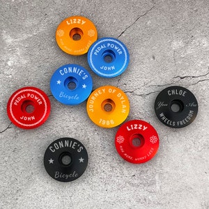 May include: Assortment of colorful bicycle stem caps with personalized text and designs.  The caps feature phrases like "Pedal Power", "Journey of Dylan", "Connie's Bicycle", and "Lizzy Ride More, Worry Less".