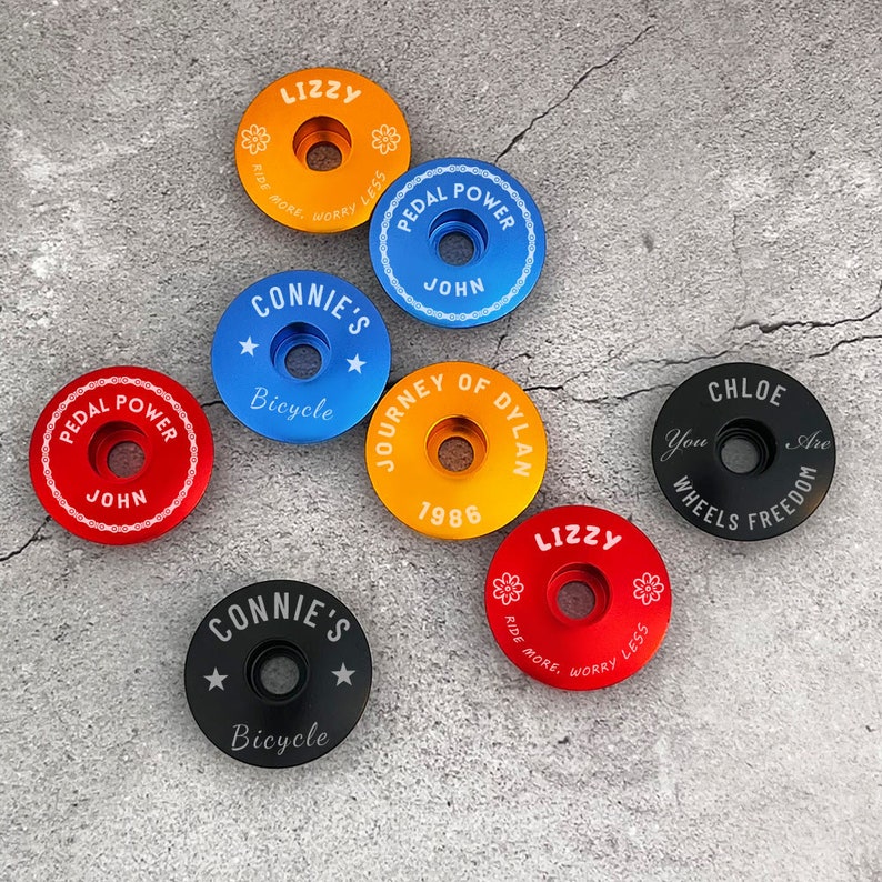 May include: Assortment of colorful bicycle stem caps with personalized text and designs.  The caps feature phrases like "Pedal Power", "Journey of Dylan", "Connie's Bicycle", and "Lizzy Ride More, Worry Less".