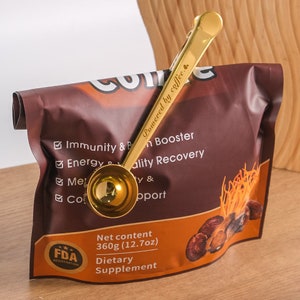 May include: Gold-colored coffee scoop with a clip attached to the handle. The clip is attached to a brown bag with the text "Powered by coffee." and "Net content 360g (12.7oz) Dietary Supplement".