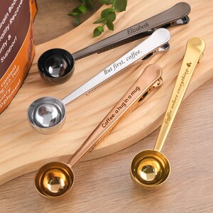 May include: Four silver coffee scoop clips with engraved sayings. The sayings are "But first, coffee", "Coffee: a hug in a mug", "Elizabeth", and "Coffee solves everything".
