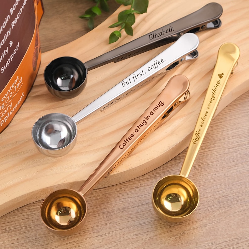 May include: Four silver coffee scoop clips with engraved sayings. The sayings are "But first, coffee", "Coffee: a hug in a mug", "Elizabeth", and "Coffee solves everything".