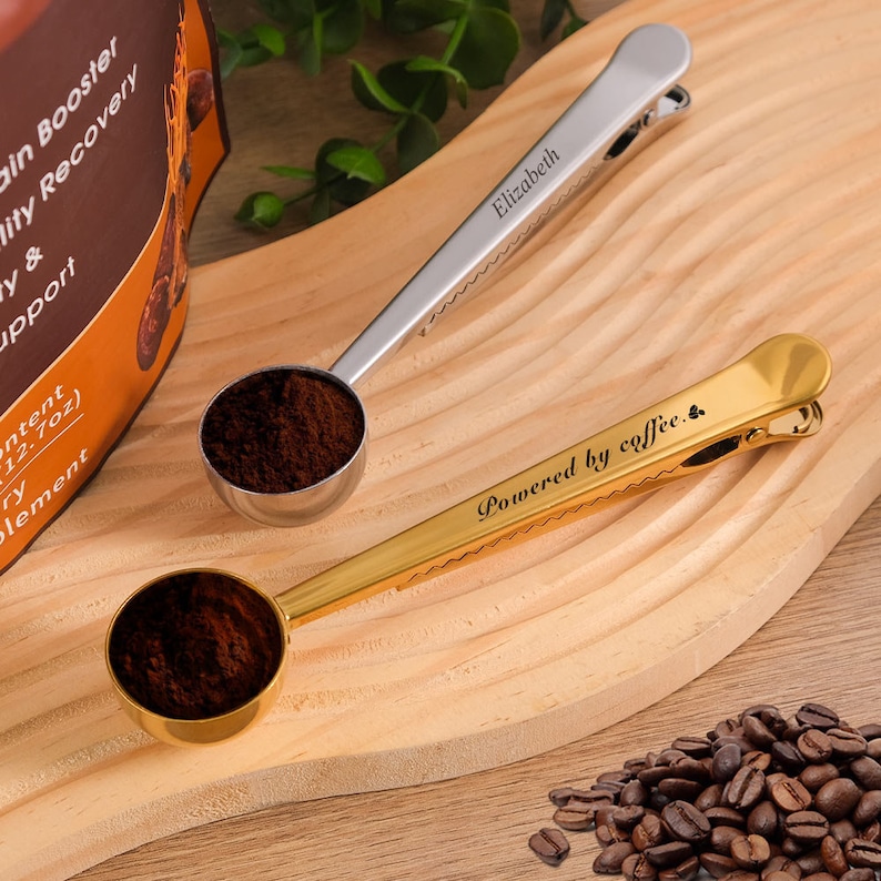 May include: Two coffee scoop clips, one gold and one silver, with personalized engravings. The gold clip says "Powered by coffee." and the silver clip says "Elizabeth".  A pile of coffee beans is in the lower right corner of the image.