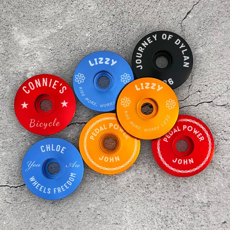 May include: A collection of six colorful bicycle stem caps with various designs and text, including "Lizzy", "Connie's Bicycle", "Journey of Dylan", "Chloe", "Pedal Power", and "John".