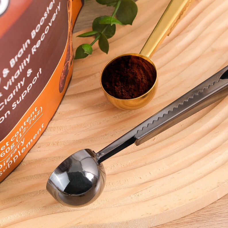 May include: Two coffee scoops, one gold and one black, with a textured handle. The scoops are filled with coffee grounds and are sitting on a wooden surface.