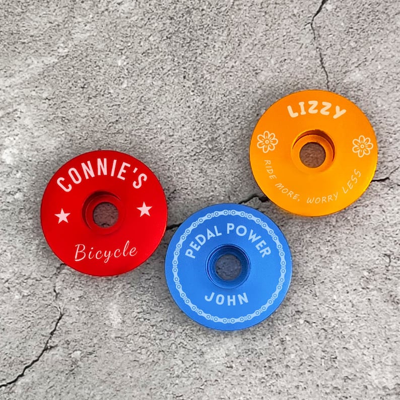 May include: Three colorful bicycle stem caps with personalized names. The red cap says "Connie's Bicycle" with white stars. The orange cap says "Lizzy Ride More Worry Less" with white flowers. The blue cap says "Pedal Power John" with a chain link design.