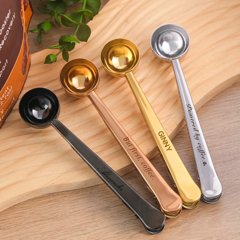 May include: Four gold, rose gold, and black coffee scoop spoons with clip handles. The spoons are engraved with the text "Powered by coffee", "GINNY", "But first, coffee", and "Alexander".