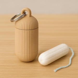 Könnte beinhalten: Ein beigefarbenes, geripptes, zylindrisches Etui mit einem silbernen Schlüsselring neben einem weißen Tampon mit hellblauer Schnur. Das Etui ist zur Aufnahme eines Tampons bestimmt. Die Artikel befinden sich auf einer hellbraunen Oberfläche.