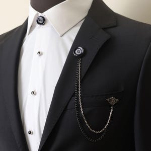 May include: A black suit jacket with a white collared shirt underneath. A black and silver chain with a decorative pin is attached to the lapel of the jacket.