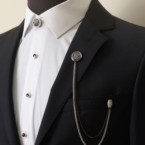 Puede incluir: Una chaqueta de traje negra con una camisa blanca de cuello debajo. Una cadena plateada con un broche circular estampado está unida a la solapa de la chaqueta. Un segundo broche plateado está unido al cuello de la camisa.