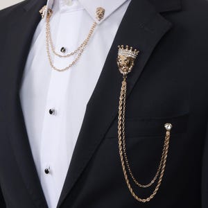 May include: A gold-toned lion head lapel pin with a crown and a chain attached. The pin is worn on a black suit jacket.