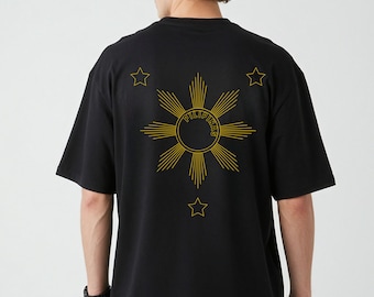 May include: Black t-shirt featuring a gold sunburst design with the word "PILIPINAS" in the center and three stars. The shirt has short sleeves and a crew neck.