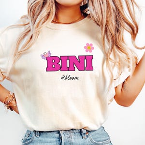 May include: Cream-colored t-shirt with the word "BINI" in bright pink block letters. Above the text is a pink flower and a butterfly. Below the text is the word "#bloom".