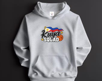 May include: A light gray hooded sweatshirt featuring a graphic print on the chest. The design includes a waving Philippine flag, a basketball, yellow stars, and the text "Kuya SQUAD" in black and white lettering. The hoodie has long sleeves.