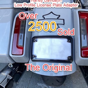 May include: A black Harley Davidson low profile license plate adapter mounted on a light gray motorcycle fender. The text reads Harley Davidson Low Profile License Plate Adapter, Over 2500 Sold, and The Original. The adapter features four mounting screws.