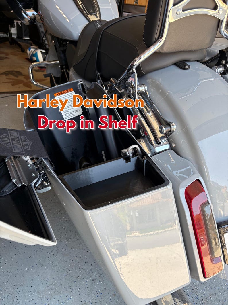 K&ouml;nnte beinhalten: Ein hellgraues Harley Davidson Motorrad mit schwarzem Sitz und einem Drop-in-Regal. Das Regal ist ge&ouml;ffnet und zeigt einen schwarzen Innenraum. Das Motorrad hat ein rotes R&uuml;cklicht und Chromakzente. Der Text "Harley Davidson Drop in Shelf" ist sichtbar.