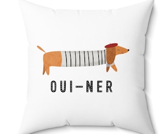 Puede incluir: Almohada blanca con un dachshund de dibujos animados que lleva una camisa a rayas y una boina roja. El texto "OUI-NER" está impreso debajo del perro.