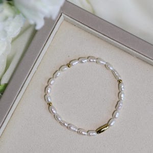 Könnte beinhalten: Ein zartes Perlenarmband mit einem goldenen Stabverschluss. Das Armband besteht aus kleinen, weißen Perlen und wird auf einem weißen Hintergrund präsentiert.