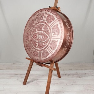 May include: A copper-colored, round decorative tray with intricate floral and geometric patterns. The tray, 78 cm in diameter and 15 cm deep, is displayed on a wooden easel. The design features a central eye-like motif and floral borders.