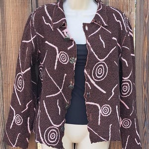 May include: A brown jacket with a white and pink abstract pattern. The jacket has a collared neckline, long sleeves, and decorative buttons. The jacket is open, revealing a black top underneath.