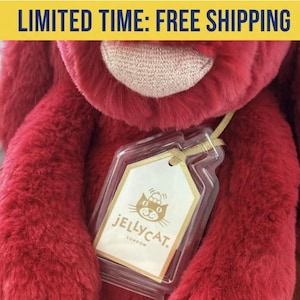 May include: A plush, red stuffed animal with a tag from Jellycat London. The tag is in a clear plastic case and features a cat logo. The stuffed animal has a soft, furry texture.