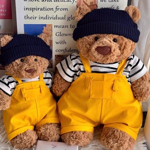 May include: Two brown plush teddy bears wearing dark navy blue knit beanies, black and white striped short-sleeve shirts, and bright yellow overalls. The bears have soft, textured fur, dark brown noses, and black eyes, presented in two sizes.