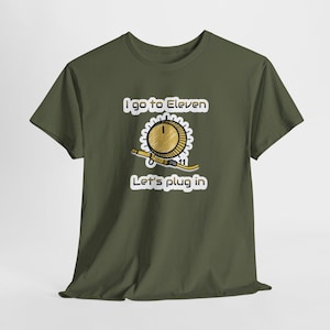 Peut inclure: T-shirt vert olive avec un motif qui dit "I go to Eleven, Let's plug in." Le motif représente un bouton doré avec le chiffre 11 et un câble. Le design est de style dessin animé.