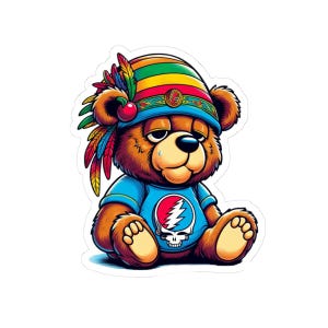 Può includere: Un adesivo con un orsetto dei cartoni animati che indossa un copricapo nativo americano colorato e una maglietta blu con un teschio e un fulmine. L'orso ha un'espressione leggermente triste.