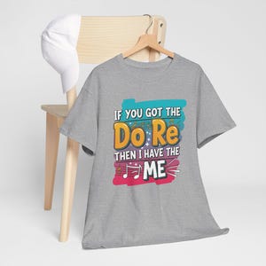 Peut inclure: Un t-shirt gris avec le texte "IF YOU GOT THE DO RE THEN I HAVE THE ME" en lettres colorées. Le t-shirt est suspendu à un cintre en bois. Une casquette de baseball blanche repose sur une chaise en bois.