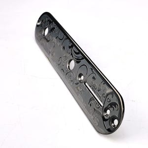 May include: A dark gray metal guitar control plate with an engraved floral design. The plate has three circular holes and a rectangular slot, with a curved, beveled edge. The decorative plate is for a guitar.