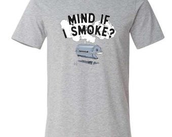 May include: A heather gray t-shirt featuring the phrase "MIND IF I SMOKE?" in black text above a cartoon illustration of a barbecue grill. The grill is silver and sits on a metal stand. The shirt has short sleeves and a crew neck.