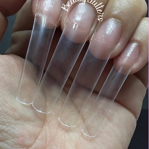 May include: Five clear plastic nail tips, also known as nail extensions, are shown on a person's hand. The tips are long and narrow, and they are designed to be applied to the natural nail to create a longer, more sculpted look.