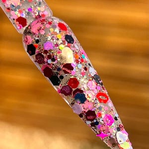 May include: A clear nail with a variety of colorful glitter, including pink, red, and gold, in various shapes and sizes. The glitter is embedded in the nail, creating a sparkling effect. The nail is tapered and pointed.
