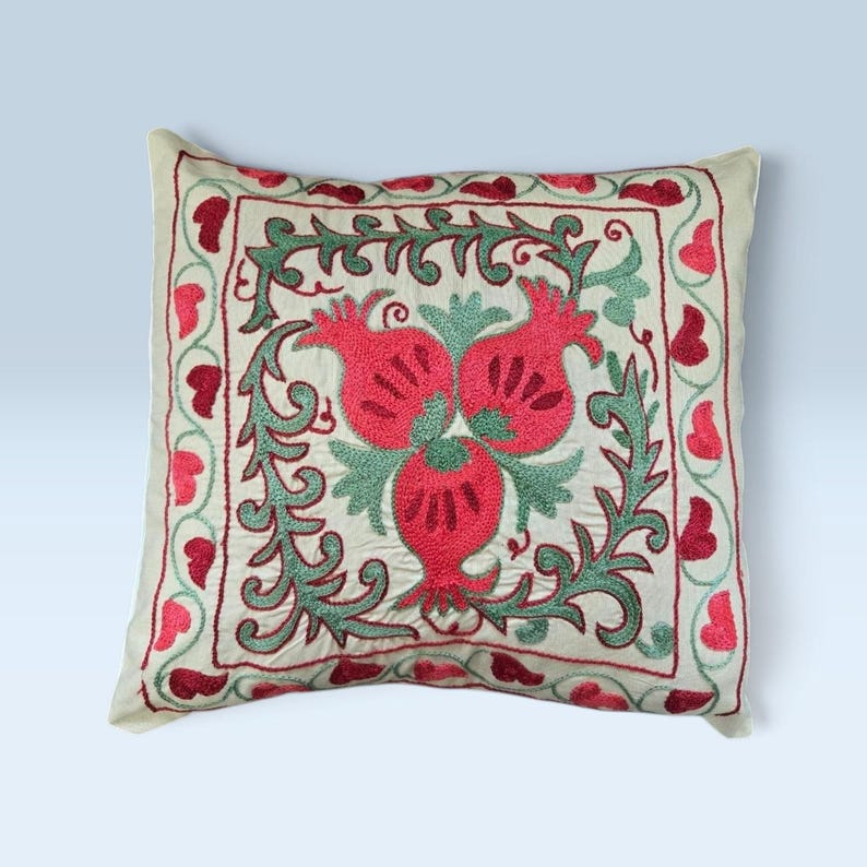 May include: A square throw cushion with an off-white fabric base featuring intricate embroidery. The design displays three stylised red pomegranate motifs surrounded by green leafy vines, enclosed within a decorative border of red heart shapes.