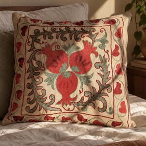 May include: A square throw cushion with a cream-coloured fabric base featuring intricate embroidery. The design displays three red pomegranate motifs surrounded by sage green leafy vines, all enclosed within a square border of red heart shapes.