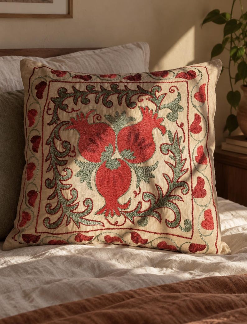 May include: A square throw cushion with a cream-coloured fabric base featuring intricate embroidery. The design displays three red pomegranate motifs surrounded by sage green leafy vines, all enclosed within a square border of red heart shapes.