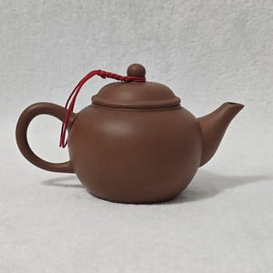 May include: A brown ceramic teapot with a curved handle and a spout. The lid has a small knob and is attached to the pot with a red string. The teapot is a matte finish and sits on a white surface.