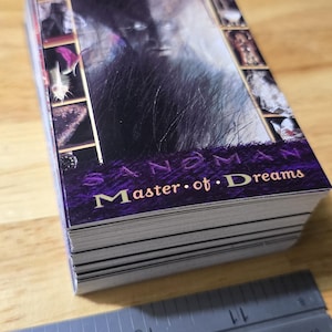 May include: A stack of cards with a dark, artistic design. The top card features a blurred image of a face and the text "Sandman Master of Dreams" in gold lettering on a purple background. A ruler is in the foreground.
