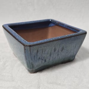 May include: A square bonsai pot with a blue glaze. The pot has a slightly textured surface and a brown interior. The pot is designed with a modern aesthetic.