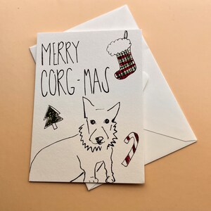 May include: A white card with a hand-drawn illustration of a corgi dog, a Christmas tree, a candy cane, and a stocking. The card says "Merry Corg-Mas".