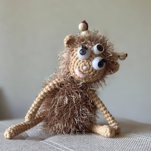Könnte beinhalten: Ein kleines, flauschiges, braunes Häkeltier mit drei Wackelaugen, einem hellbeigen Gesicht und zwei Antennen, die mit kleinen Holzperlen verziert sind. Der Körper ist mit zotteligem, strukturiertem Garn bedeckt und hat dünne, bewegliche Häkelarme und -beine.