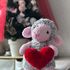 May include: A handmade crochet lamb plushie in gray, pink, and red. The lamb is holding a red heart. The background is blurred, but a Christmas tree is visible. This soft toy is perfect for Valentine's Day or as a gift.